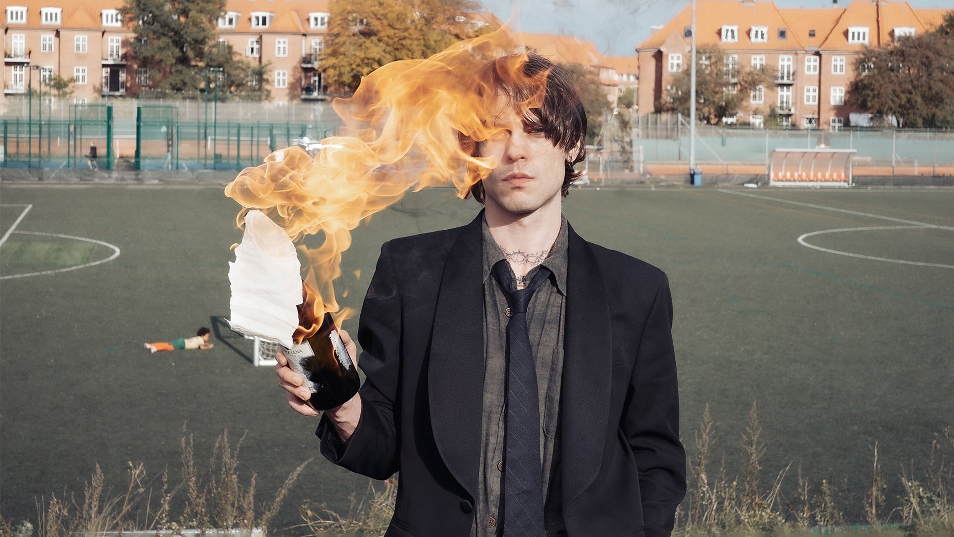 Foto für die Veranstaltung ‚ELIAS RØNNENFELT‘ mit dem Künstler, der ein Papier in einer Flasche verbrennt, vor dem Hintergrund eines Fußballfeldes
