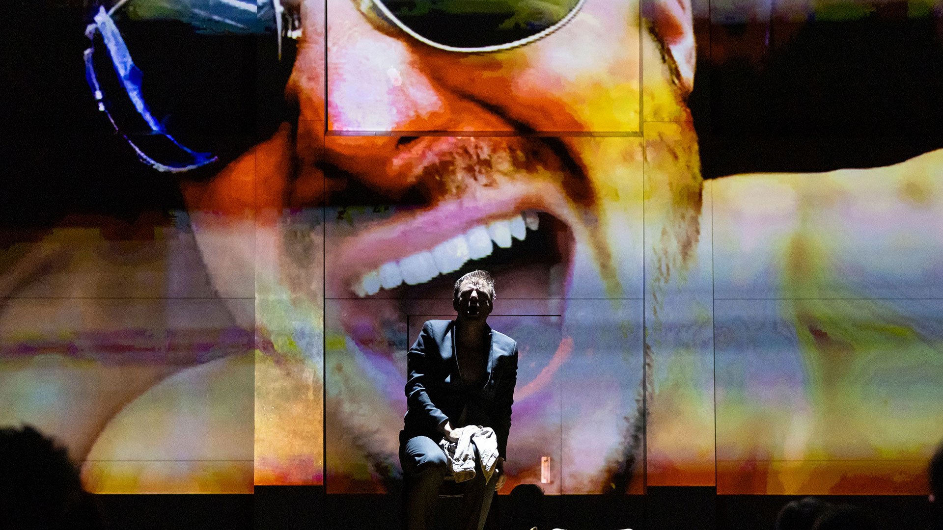 Bühnenszene mit einem sitzenden Mann im Vordergrund, hinter ihm eine große farbige Projektion eines Gesichts mit Sonnenbrille – Theatermotiv.