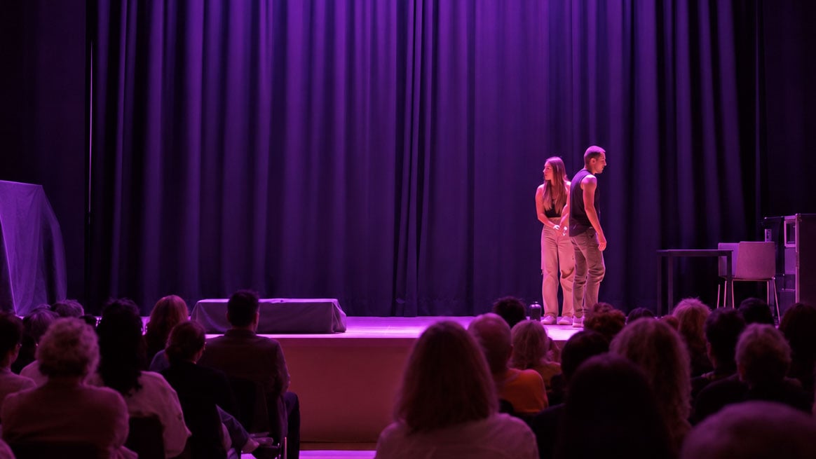 Zwei Personen stehen auf einer Theaterbühne vor violett beleuchtetem Vorhang und einem sitzenden Publikum.