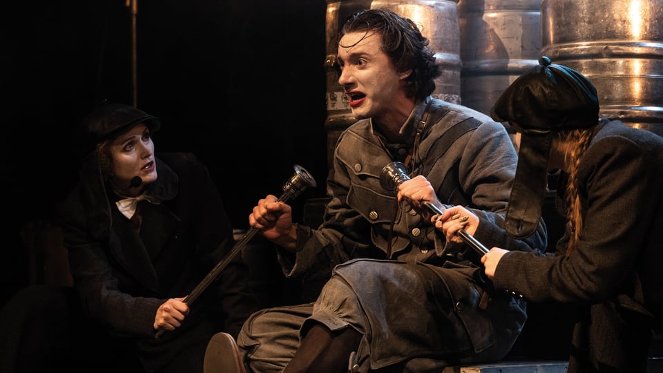 Theaterszene mit drei Personen auf einer Bühne. Eine Person sitzt in einem historischen Kostüm im Mittelpunkt und hält zwei Stäbe in den Händen. Zwei weitere Personen in dunklen Kostümen sitzen oder knien daneben. Im Hintergrund stehen mehrere große Metallfässer, die von warmem Bühnenlicht angestrahlt werden.