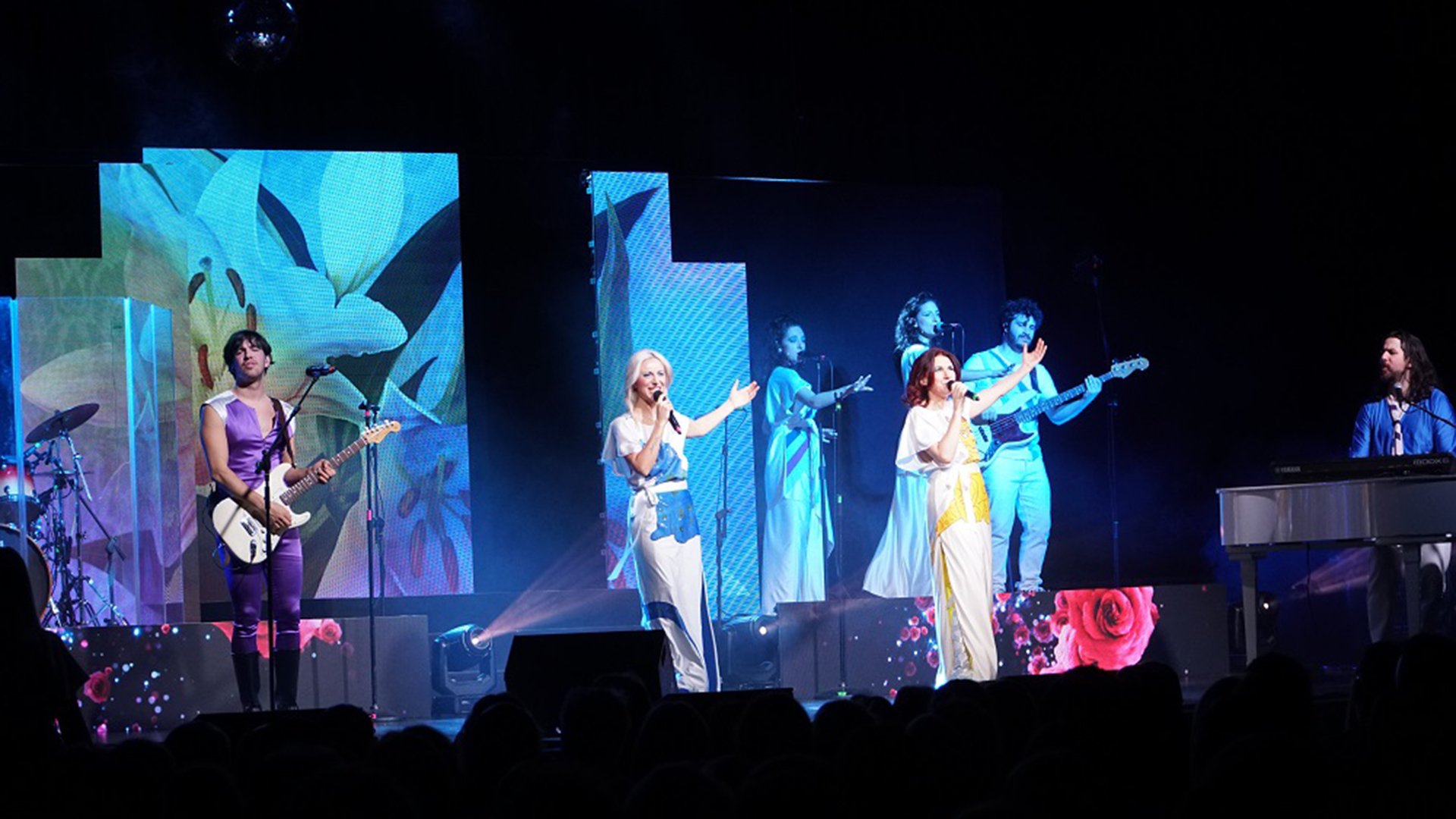 Band auf Bühne im ABBA-Stil mit Sängerinnen, Musikern und blauem Bühnenlicht