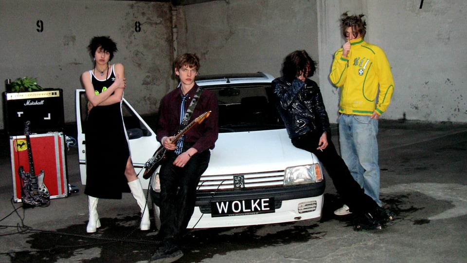 Die vier Bandmitglieder in einer Halle vor einem weißen Auto