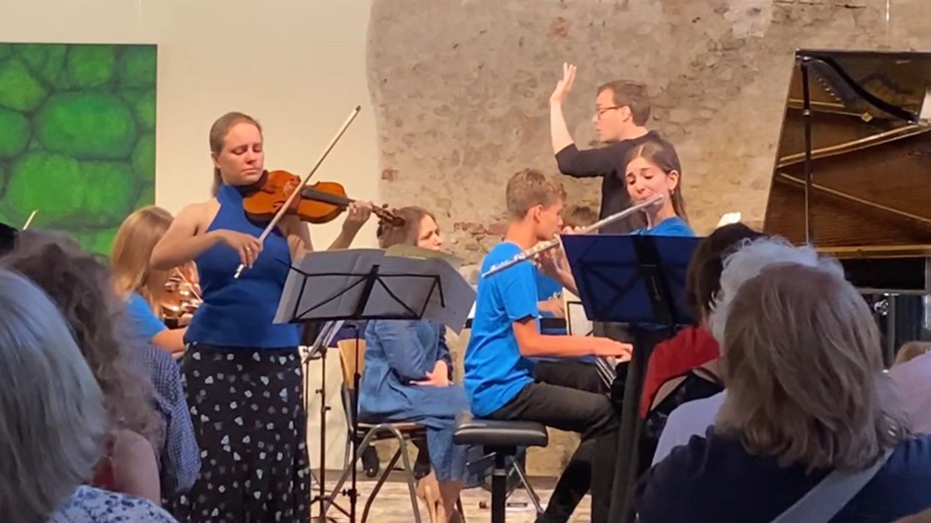 Veranstaltungsfoto eines klassischen Konzerts mit jungen Musikerinnen und Musikern: Mehrere Personen musizieren auf einer kleinen Bühne vor Publikum. Im Vordergrund spielt eine Person Geige, rechts daneben spielt eine weitere Person Querflöte, begleitet von einer Person am Klavier. Im Hintergrund steht eine dirigierende Person. Notenständer sind aufgestellt, das Publikum ist teilweise im Vordergrund zu sehen. Die Aufführung findet in einem Innenraum mit heller Wand, sichtbarer Mauerstruktur und warmer Beleuchtung statt und vermittelt die Atmosphäre eines Live‑Kammer‑ oder Nachwuchskonzerts.