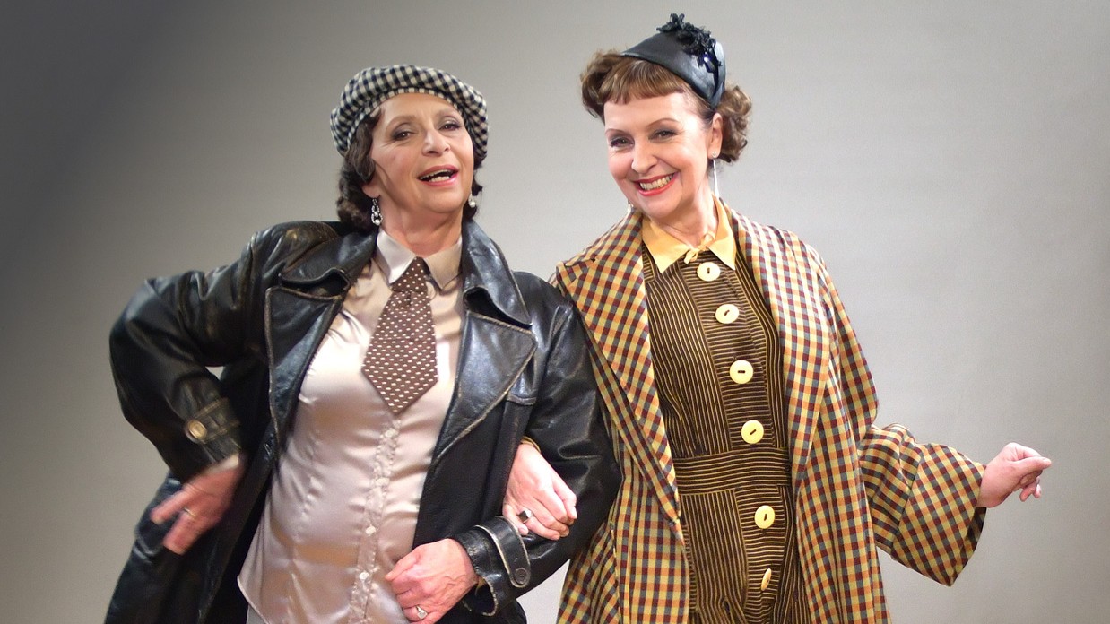Susanne Brantl und Conny Glogger in historisch inspirierter Kleidung auf einer Bühne. Eine Frau trägt Lederjacke, Krawatte und karierte Mütze, die andere einen karierten Mantel mit schwarzem Hut. Beide haken sich unter und lächeln.