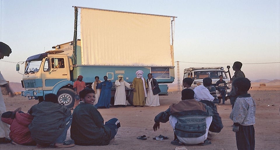 Mehrere Personen sitzend und stehend in der Wüste, im Hintergrund ein Lkw mit Leinwand