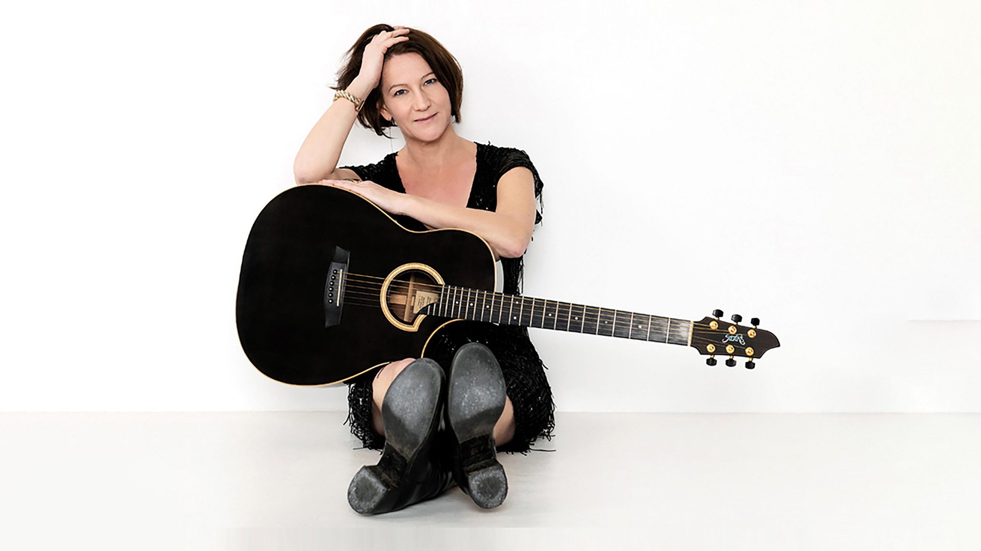 Ein Foto der Künstlerin Mira Cardui, die auf dem Boden sitzt, mit einer Gitarre vor weißem Hintergrund