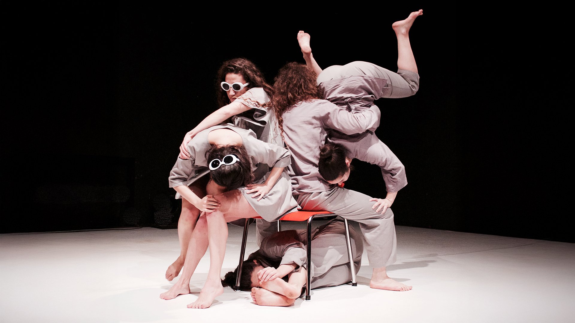 Bühnenfoto aus einem zeitgenössischen Tanzstück: Fünf Performer*innen sind barfuß auf einer runden, hellen Bühne arrangiert und formen gemeinsam eine skulpturale Gruppe um einen Stuhl. Die Körper liegen, sitzen und stützen sich aufeinander. Alle tragen helle, einfarbige Kostüme sowie runde, weiße Brillen. Schwarzer Bühnenhintergrund, fokussierte Beleuchtung auf die Tanzszene.