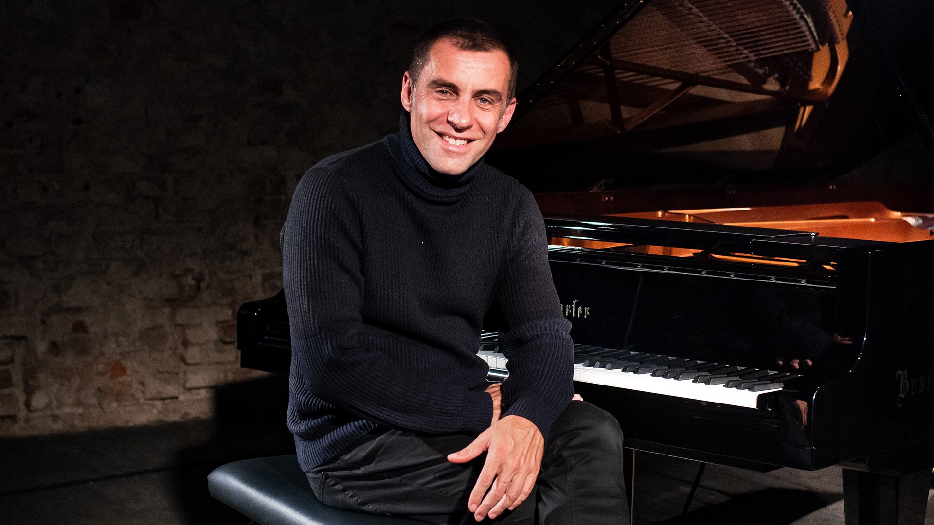 Pianist sitzt lächelnd an einem Flügel auf einer Bühne, Porträtfoto in dunkler Konzertatmosphäre.
