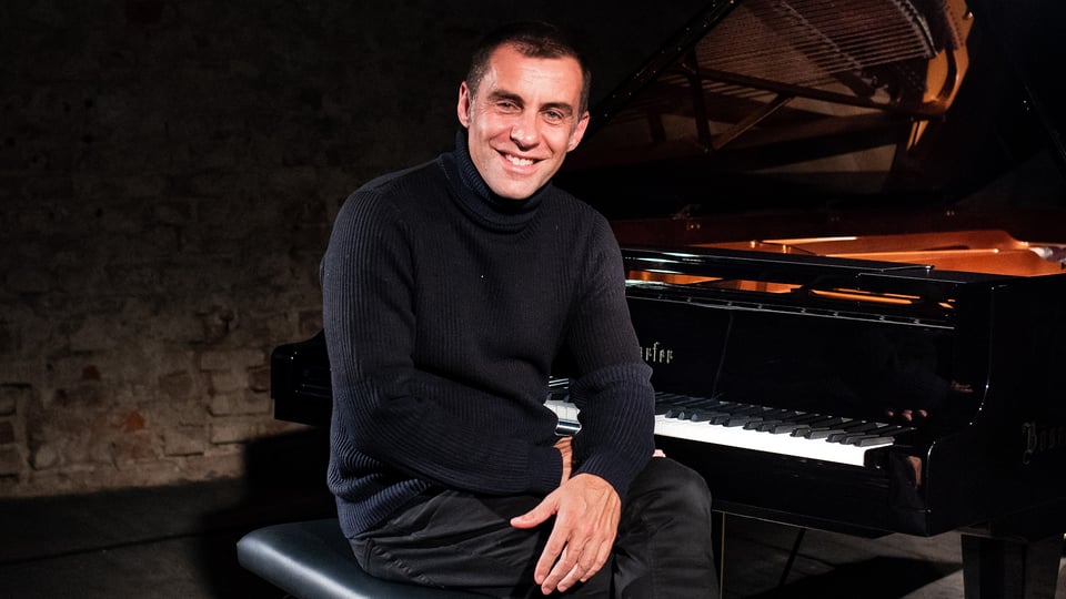 Pianist sitzt lächelnd an einem Flügel auf einer Bühne, Porträtfoto in dunkler Konzertatmosphäre.