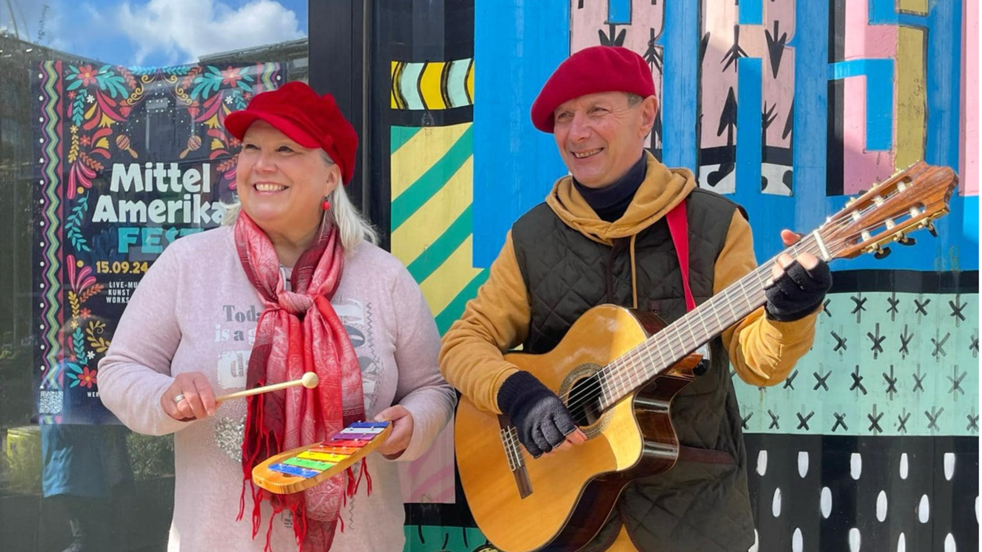 Foto mit zwei Personen vor bunter Wand mit grafischen Mustern und Plakat Mittel Amerika Fest eine Person mit Xylofon die andere mit Gitarre beide mit roten Mützen