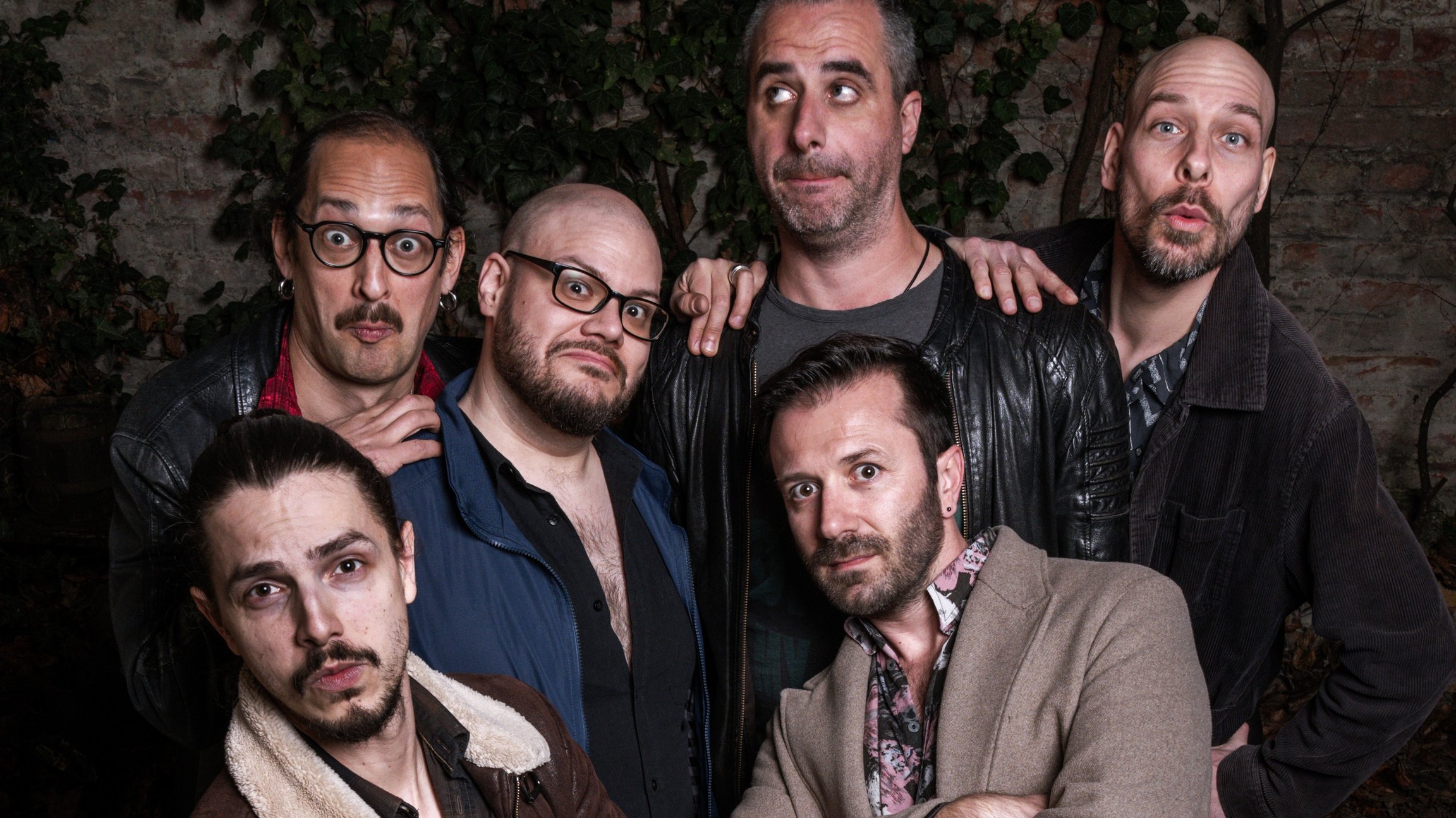 Bandfoto eines Sextetts mit sechs Personen in dunkler Freizeitkleidung, die dicht beieinander in einem Innenhof vor einer bewachsenen Backsteinwand stehen, einige mit verschränkten Armen und unterschiedliche Posen zur Kamera einnehmen.
