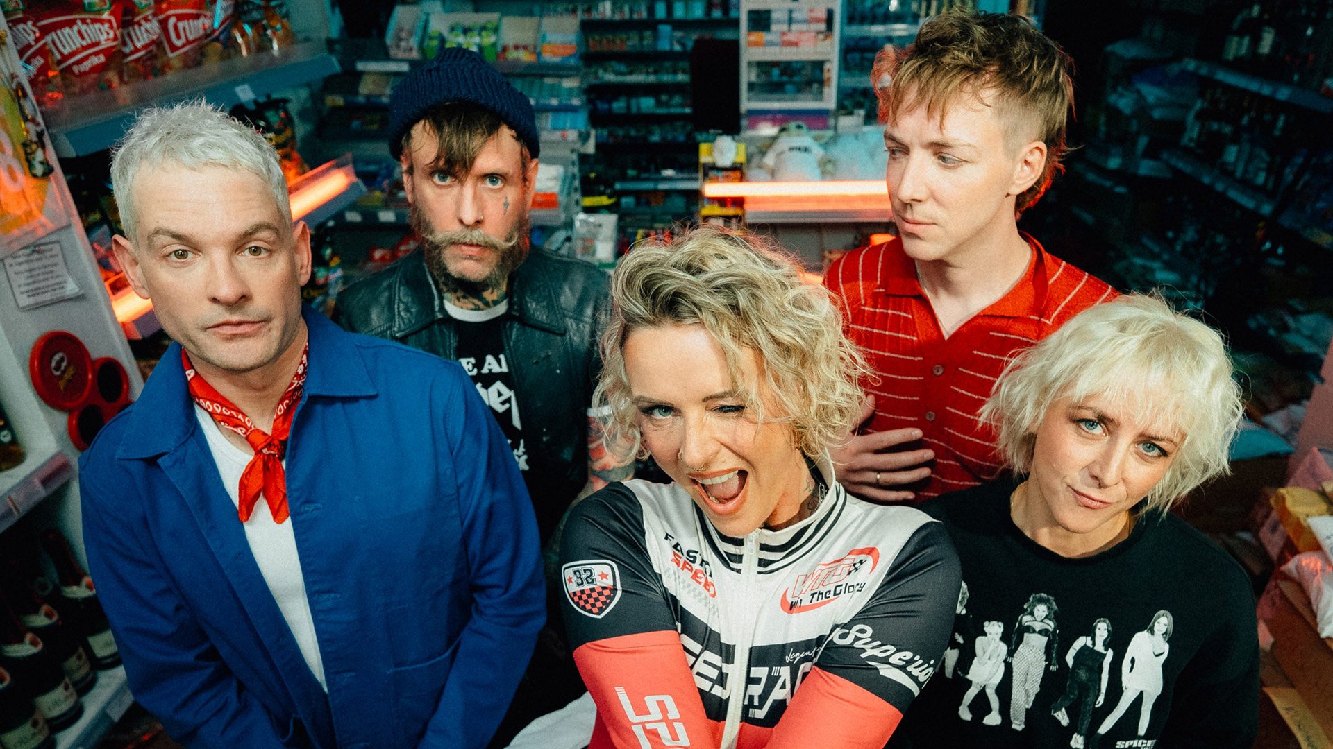 Bandfoto von Jennifer Rostock: Fünf Personen stehen in einem Kiosk zwischen Regalen und blicken in die Kamera. Die Szene ist farbenfroh ausgeleuchtet und wirkt modern und dynamisch.