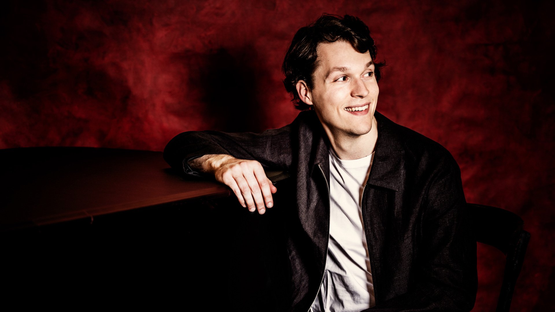 Porträt des Pianisten Lukas Sternath am Flügel vor rotem Hintergrund.