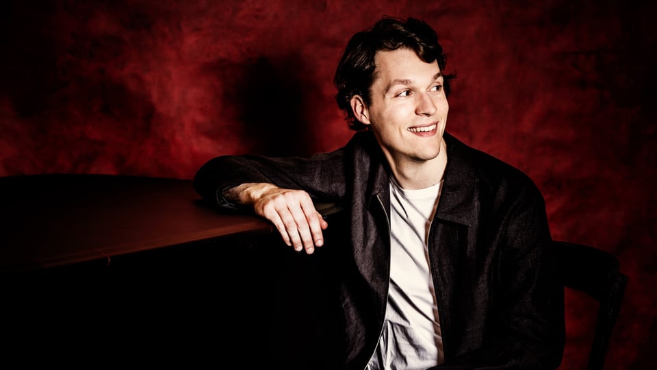 Porträt des Pianisten Lukas Sternath am Flügel vor rotem Hintergrund.