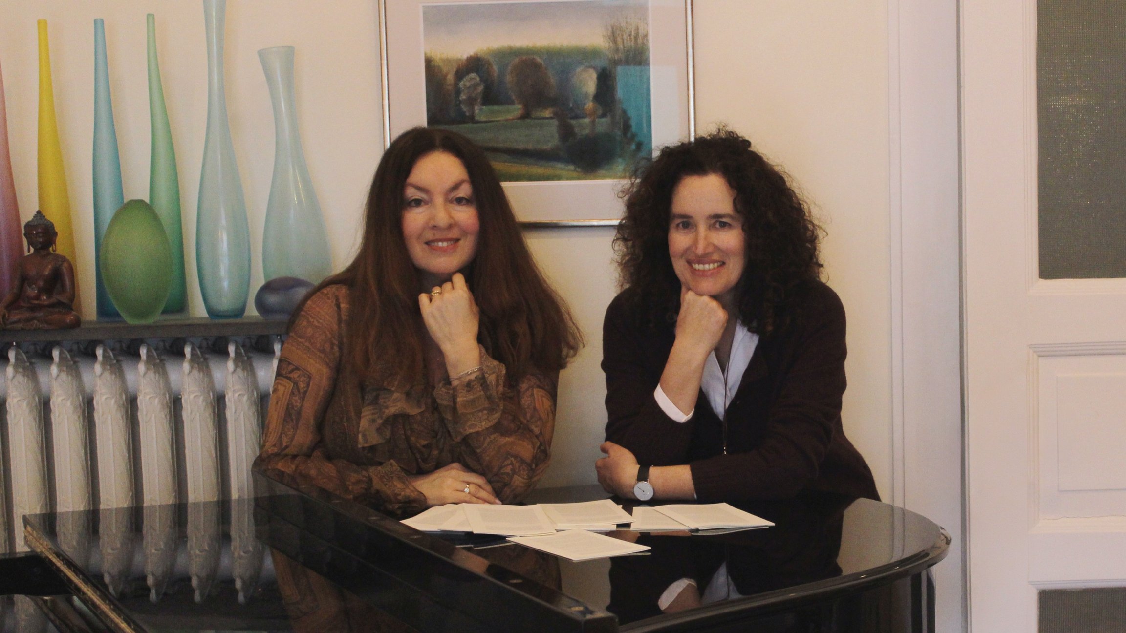 Zwei Frauen sitzen an einem Flügel in einem Innenraum, lehnen sich auf den Tisch mit Notenblättern, im Hintergrund stehen Vasen und ein gerahmtes Bild an der Wand.