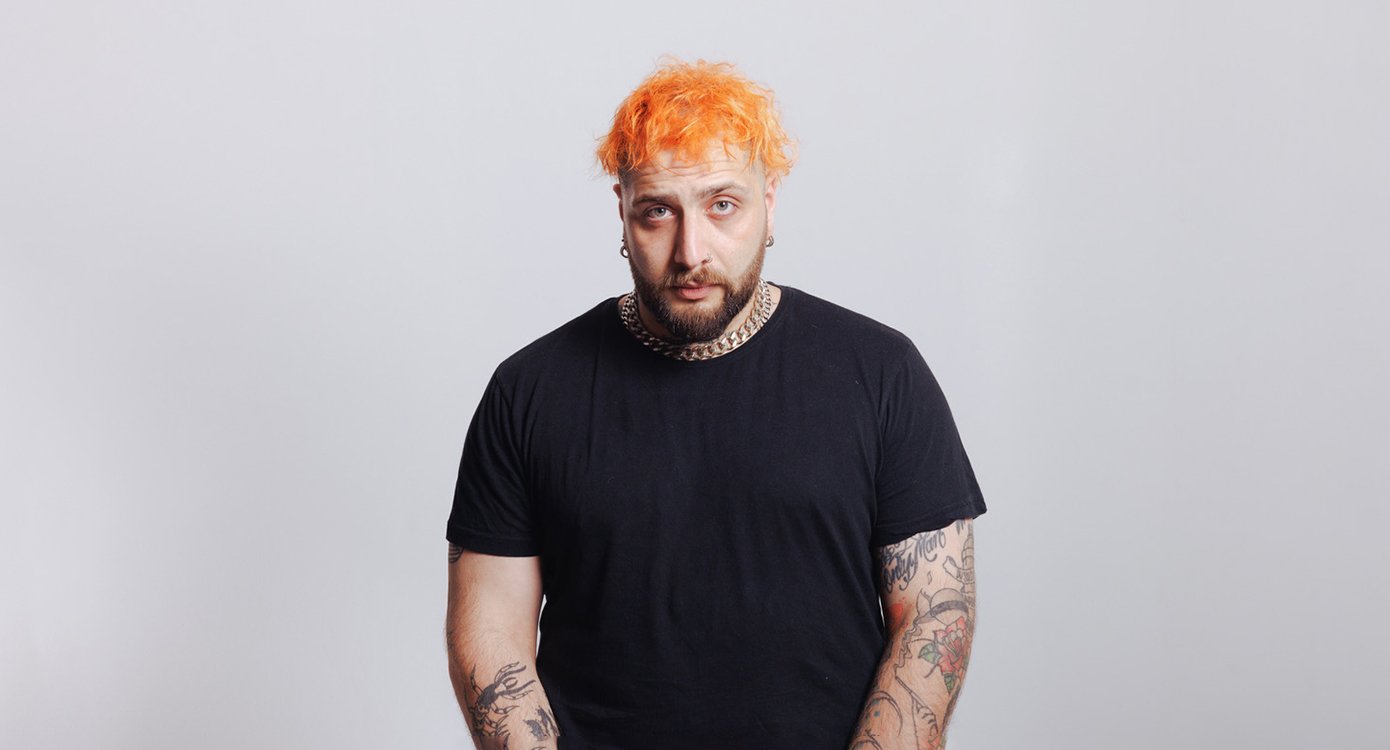 Ein Foto des Künstlers mit orangefarbenem Haar und schwarzem T-Shirt vor grauem Hintergrund