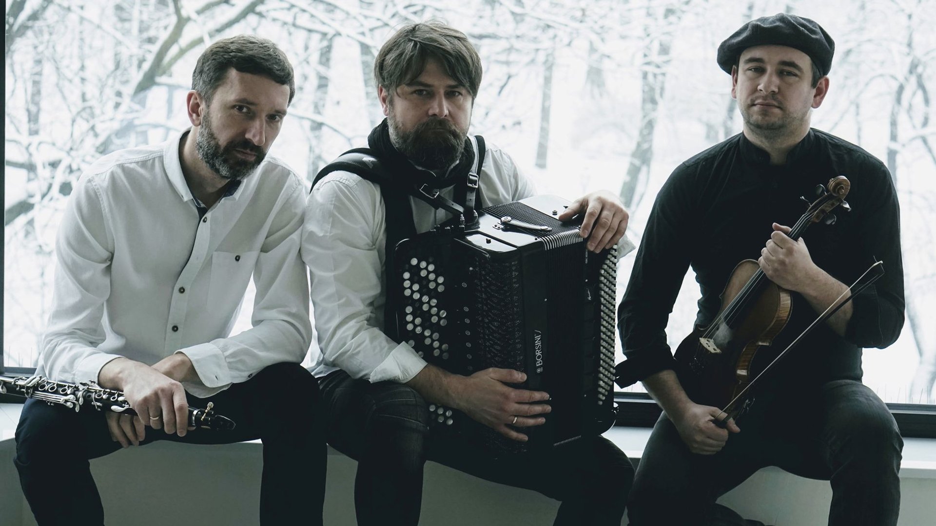 rio m.o.l.o vor einem großen Fenster mit winterlicher Landschaft. Die drei Musiker tragen schwarze und weiße Kleidung und halten Klarinette, Knopfakkordeon und Violine.