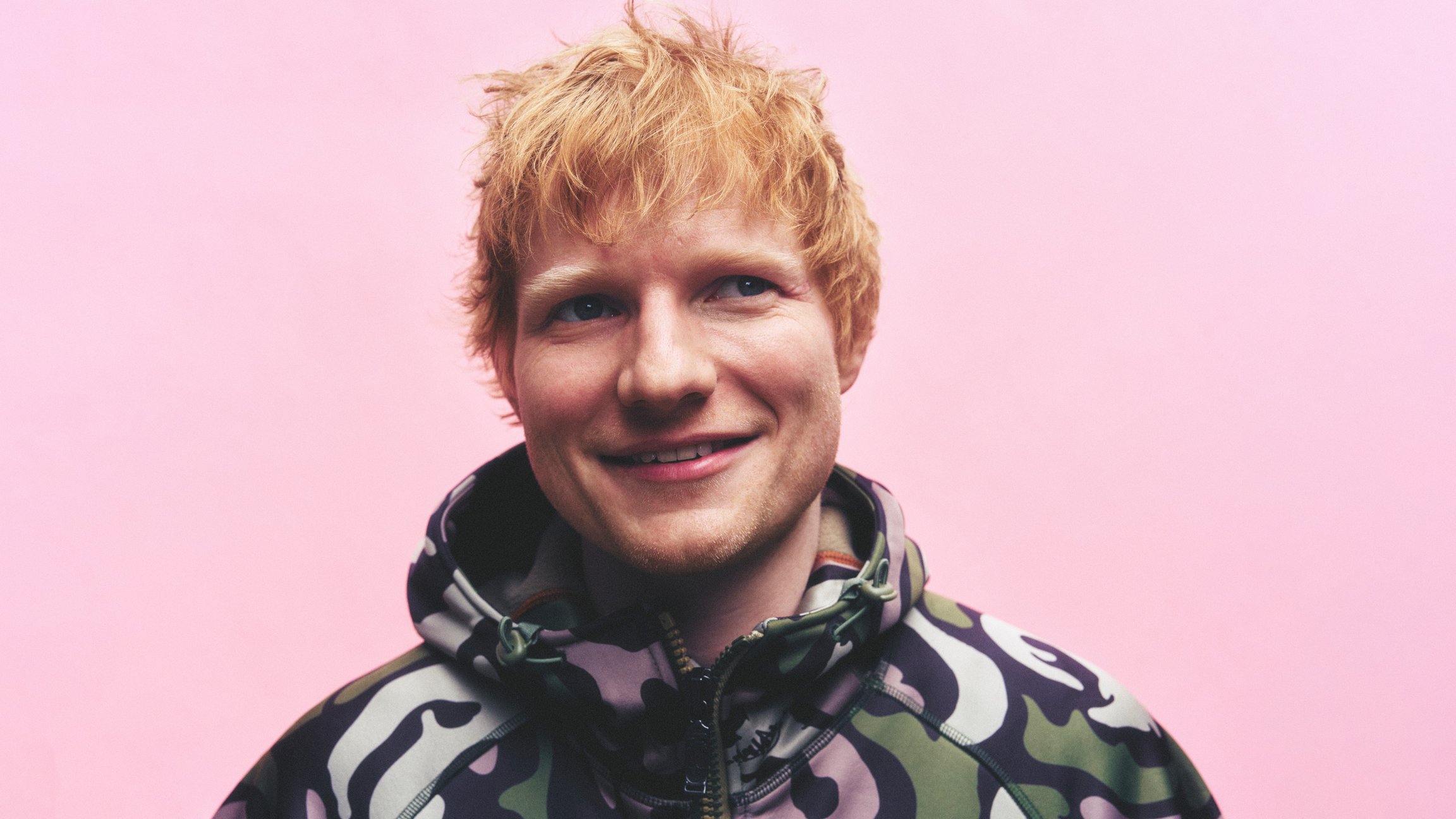 Porträt von Ed Sheeran während er lächelt vor einem rosanen Hintergrund