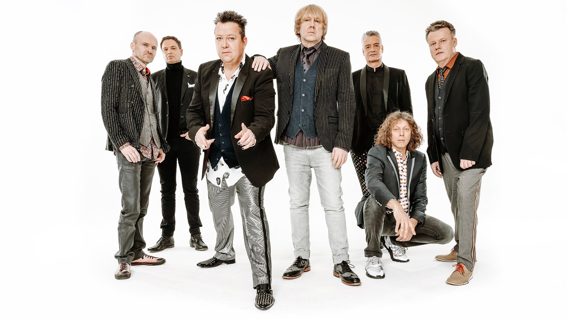 Gruppenfoto der Band Die Prinzen: sieben Musiker stehen und sitzen vor weißem Hintergrund, tragen dunkle Sakkos und Hemden in unterschiedlichen Mustern und Posen.