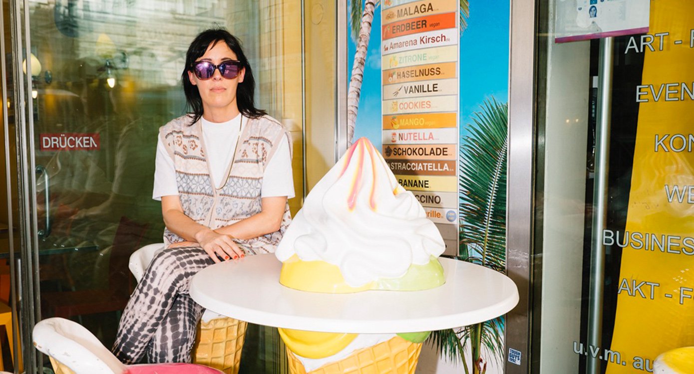 Künstlerin mit Sonnenbrille in einer Eisdiele mit übergroßem Eis als Tisch sitzend