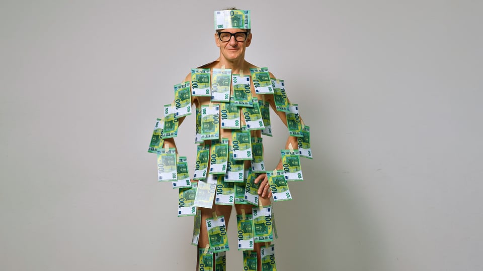 Person mit Brille steht vor hellem Hintergrund und ist am ganzen Körper mit 100‑Euro‑Banknoten beklebt