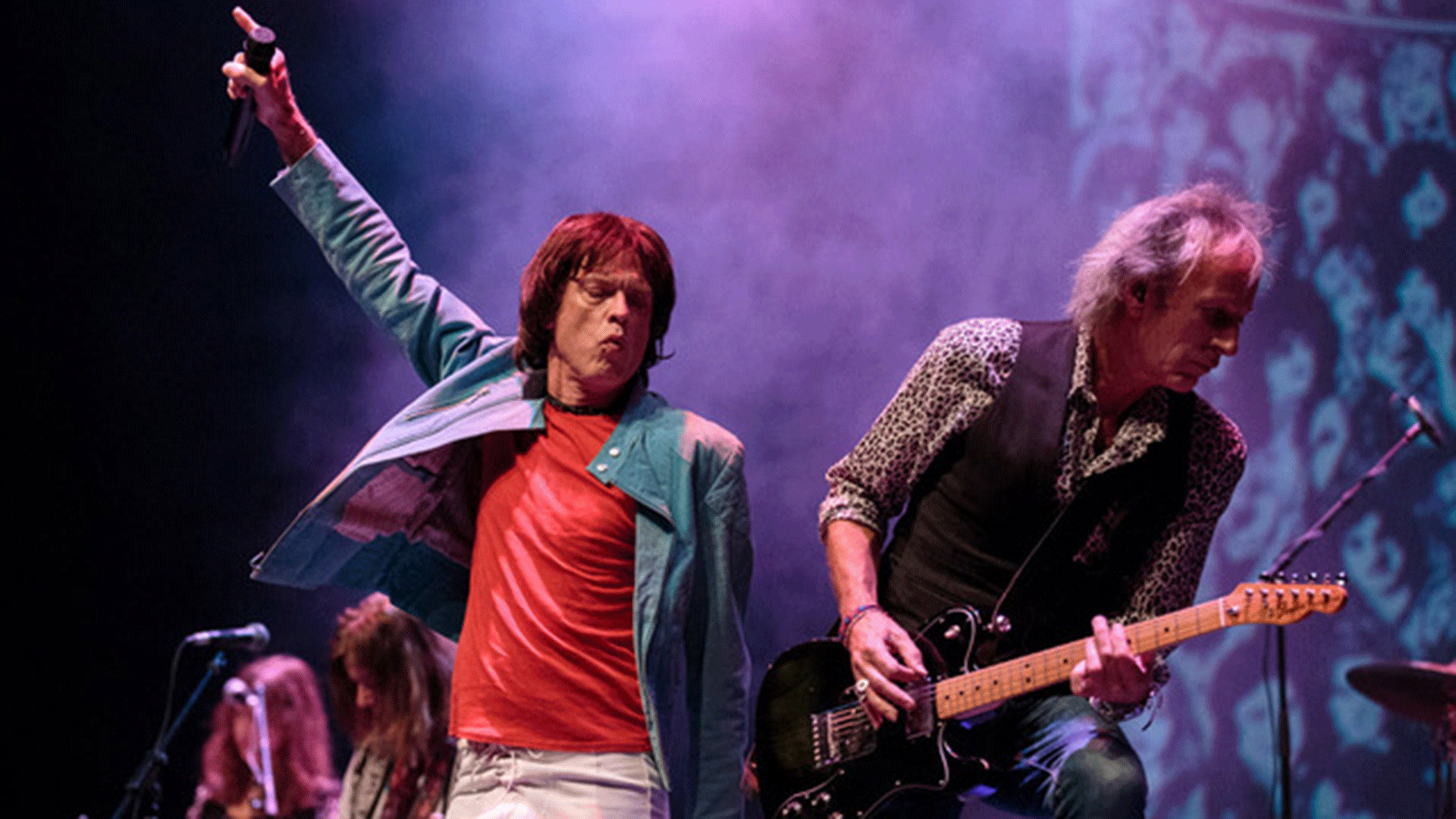 Liveauftritt der Rolling Stones auf einer Konzertbühne. Eine Person im roten Shirt hält ein Mikrofon in die Höhe, während eine weitere Person E Gitarre spielt. Im Hintergrund sind weitere Bandmitglieder sowie farbige Bühnenbeleuchtung und Nebeleffekte zu sehen.