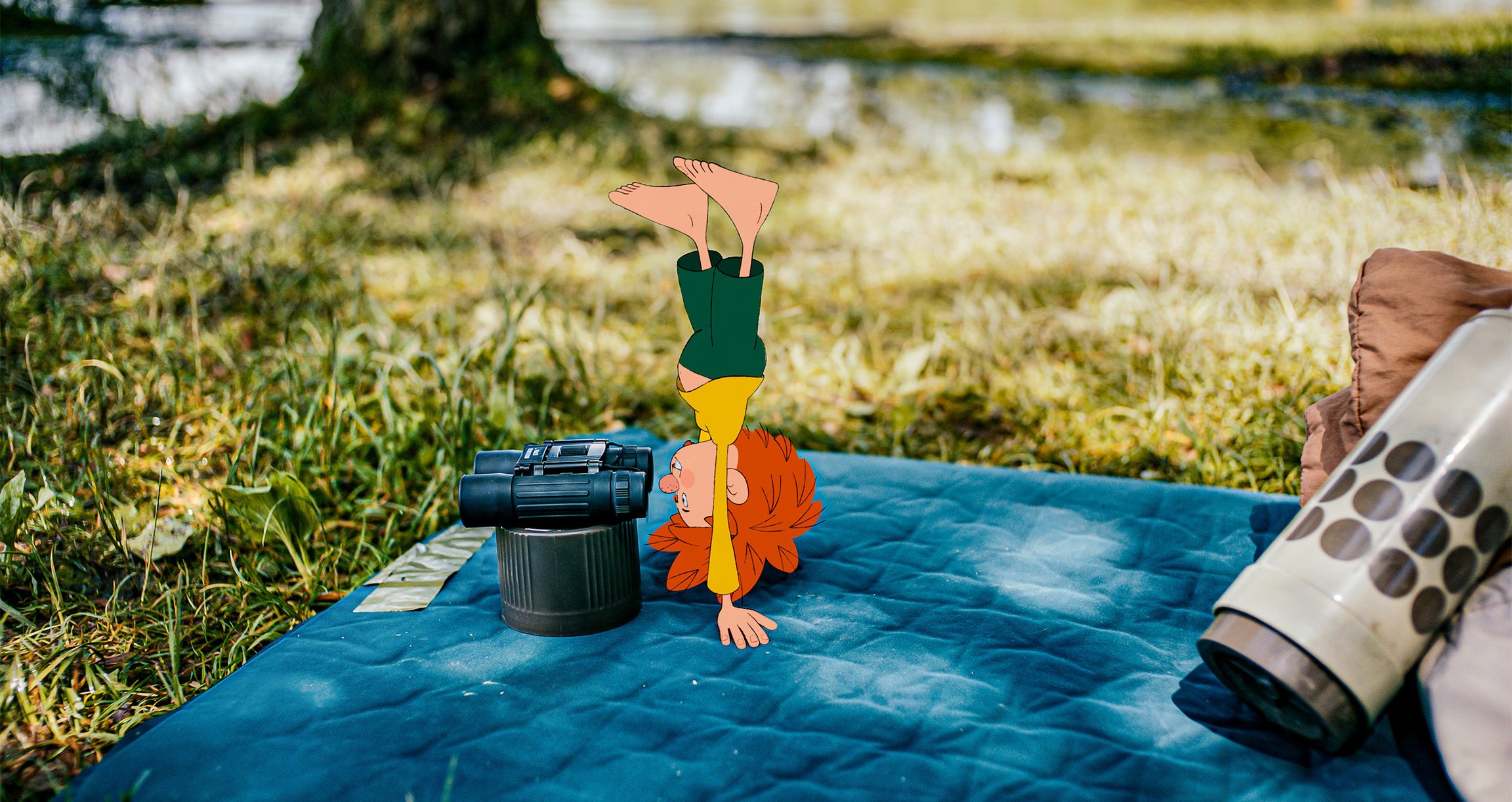 Das Pumuckl guckt im Handstand durch ein Fernglas auf einer Decke auf einer Wiese