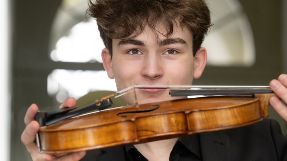 Der Künstler Erik Maier in einem schwarzen Hemd, im Vordergrund sein Streichinstrument, im Hintergrund ein rundes Glasfenster mit unscharfen Konturen