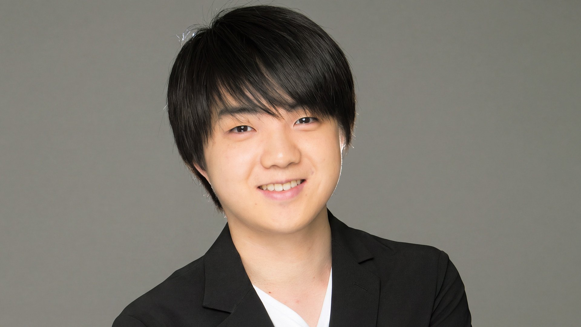 En Portrait des Künstlers Mao Fujita lächelnd in schwarzem Sakko vor grauem Hintergrund