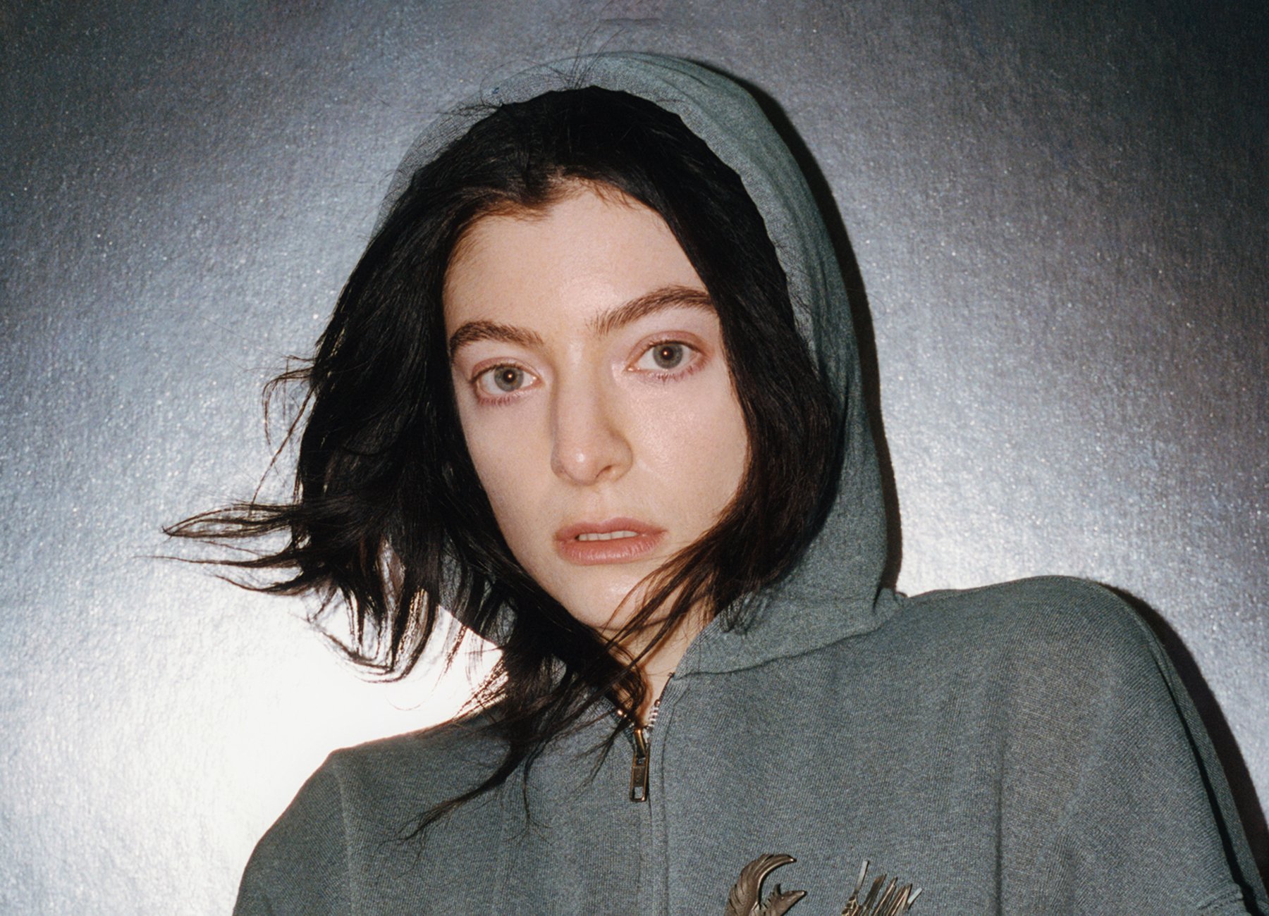 Lorde steht mit einer grauen Zip-Jacke vor einer grauen Wand und guckt mit Kapuze in die Kamera