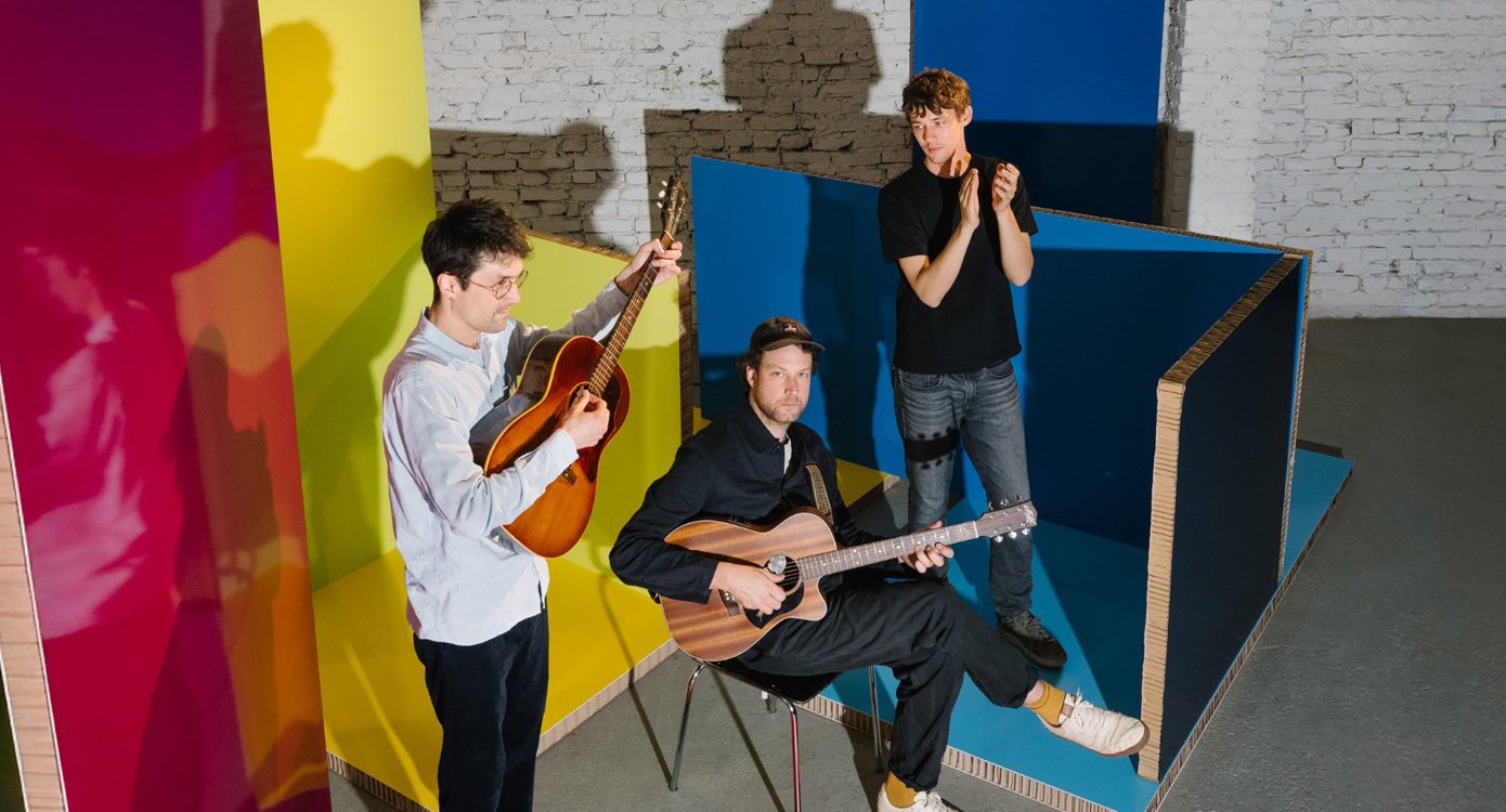 Die Band steht in einem Raum und spielt. Zwei haben eine Gitarre und einer klatscht in die Hände.