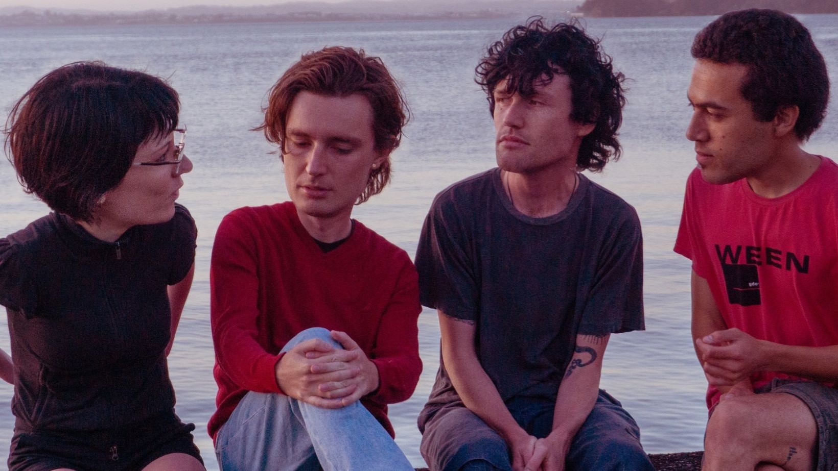 Die vier Mitglieder der band Ringlets sitzen nebeneinander an einem Gewässer und tauschen sich aus, im Hintergrund ist Wasser und eine entfernte Uferlinie zu sehen