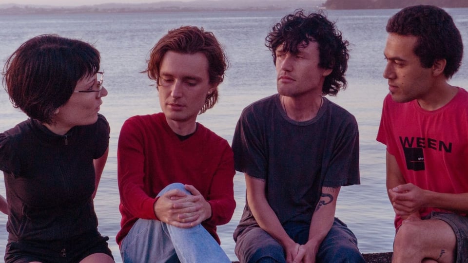 Die vier Mitglieder der band Ringlets sitzen nebeneinander an einem Gewässer und tauschen sich aus, im Hintergrund ist Wasser und eine entfernte Uferlinie zu sehen