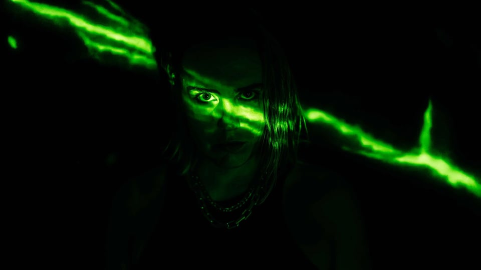 Person frontal im Dunkeln, nur Gesicht und Oberkörper sichtbar, neon‑grüne Lichtstreifen verlaufen diagonal über Augen und Haar vor schwarzem Hintergrund