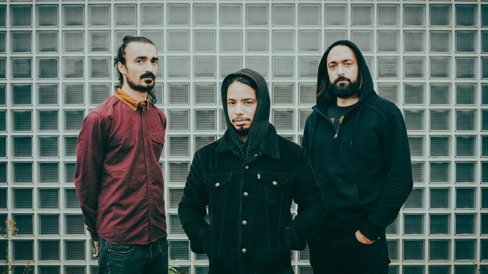 Bandfoto von SLIFT vor einer Wand aus Glasbausteinen. Die Personen tragen dunkle Freizeitkleidung mit Kapuzenjacken und Hemd und stehen in lockerer Formation nebeneinander vor der geometrischen Fassadenstruktur.
