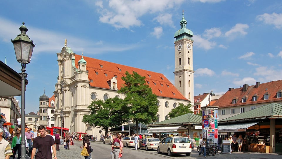 Stadtansicht von München am Viktualienmarkt: Blick auf die Heiliggeistkirche mit hellem Kirchturm und rotem Ziegeldach, umgeben von Marktständen, Passantinnen und Passanten sowie parkenden Fahrzeugen; belebte Innenstadt bei blauem Himmel.