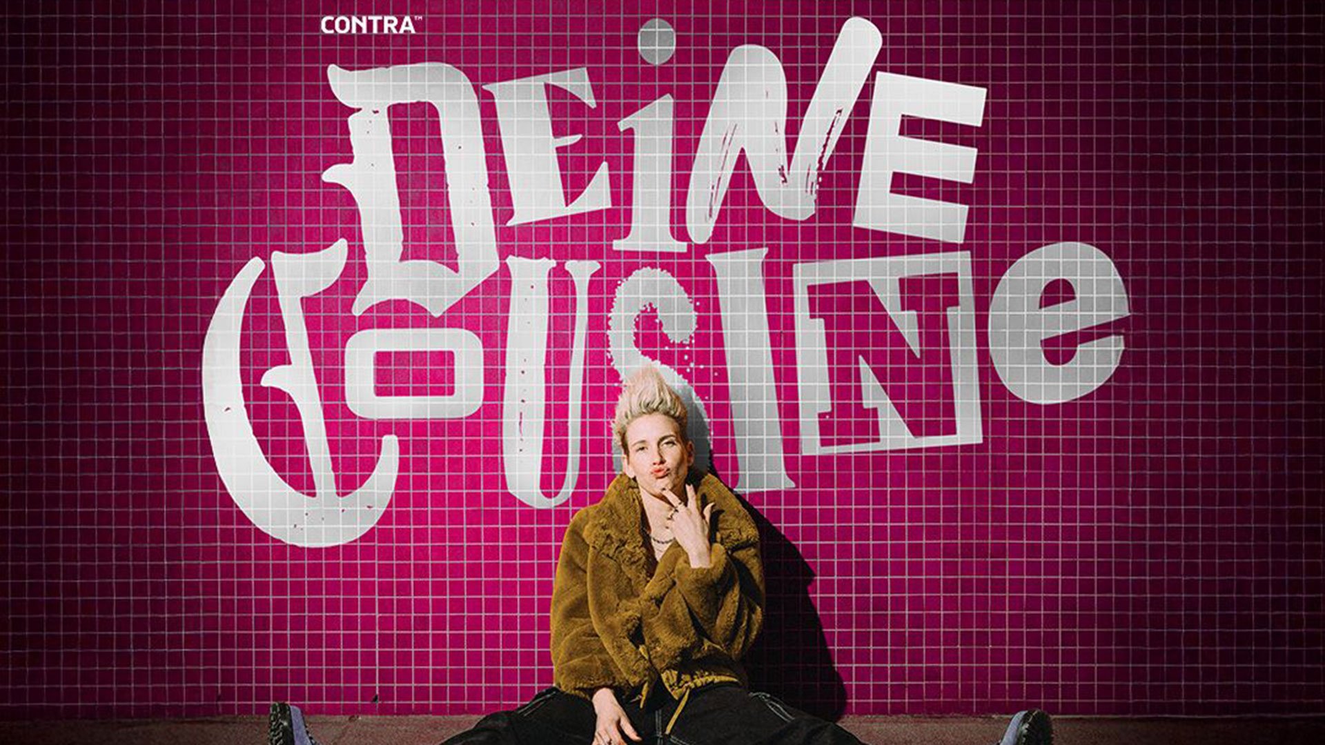 Eventfoto „DEINE COUSINE – LIVE 2026 – Ich weiß, wie dieser Abend endet!“ mit der Künstlerin sitzend auf dem Boden vor rosa kariertem Hintergrund mit Künstlernamen.