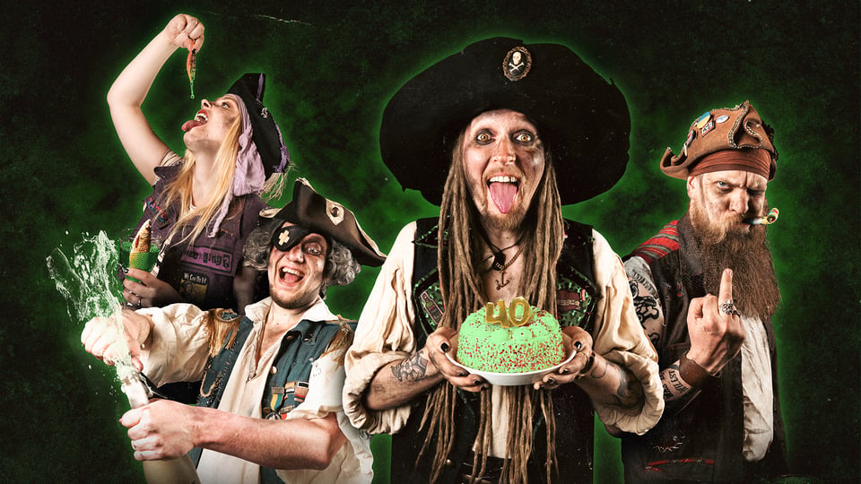 Vier Personen in Piratenkostümen vor grünem Hintergrund, eine Person hält eine Torte mit 40, andere mit Flasche und bunten Requisiten, Gruppenmotiv für Party‑Konzert