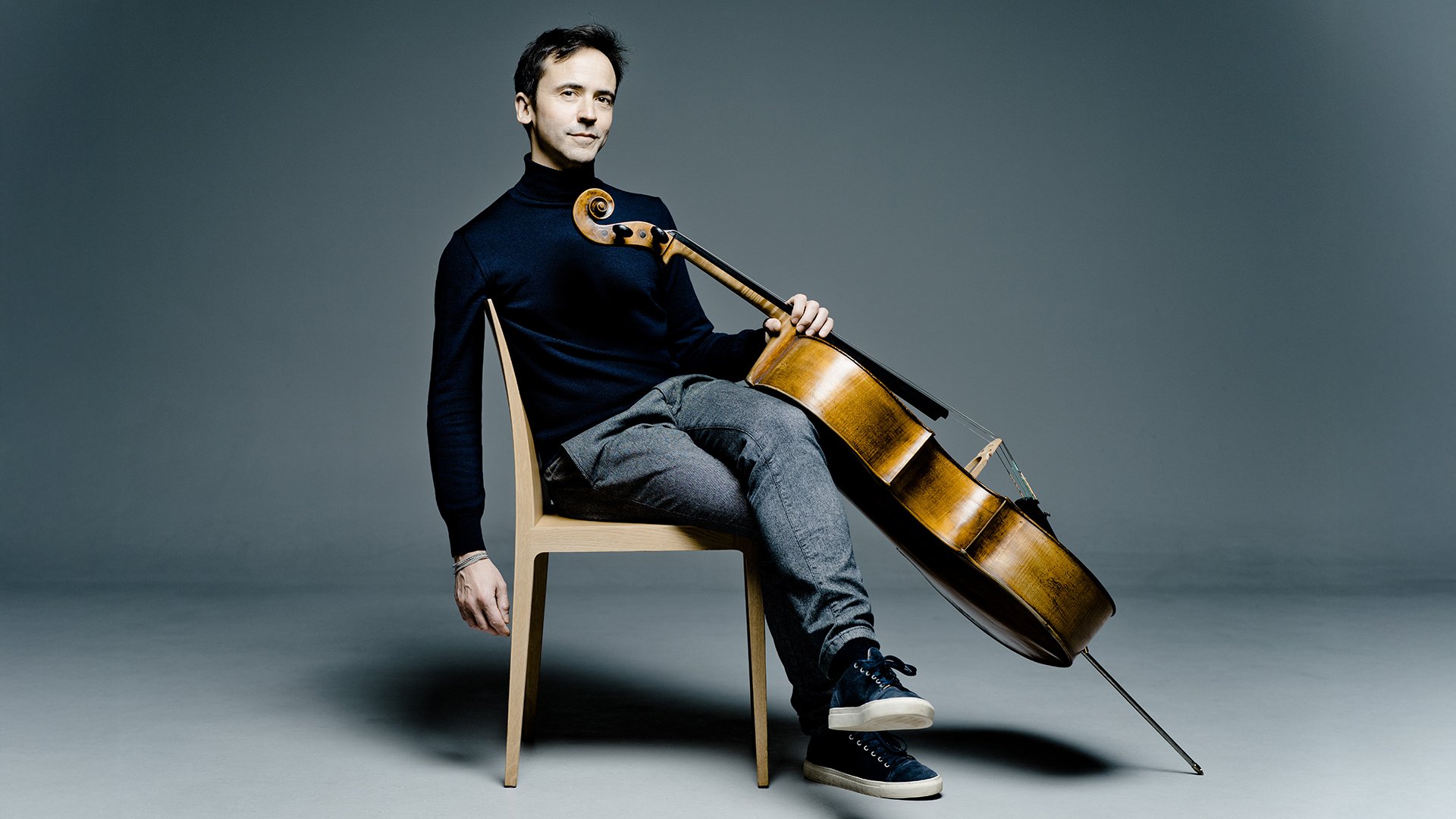 Jean Guihen Queyras sitzt auf einem Stuhl und hält ein Cello vor neutralem Hintergrund.