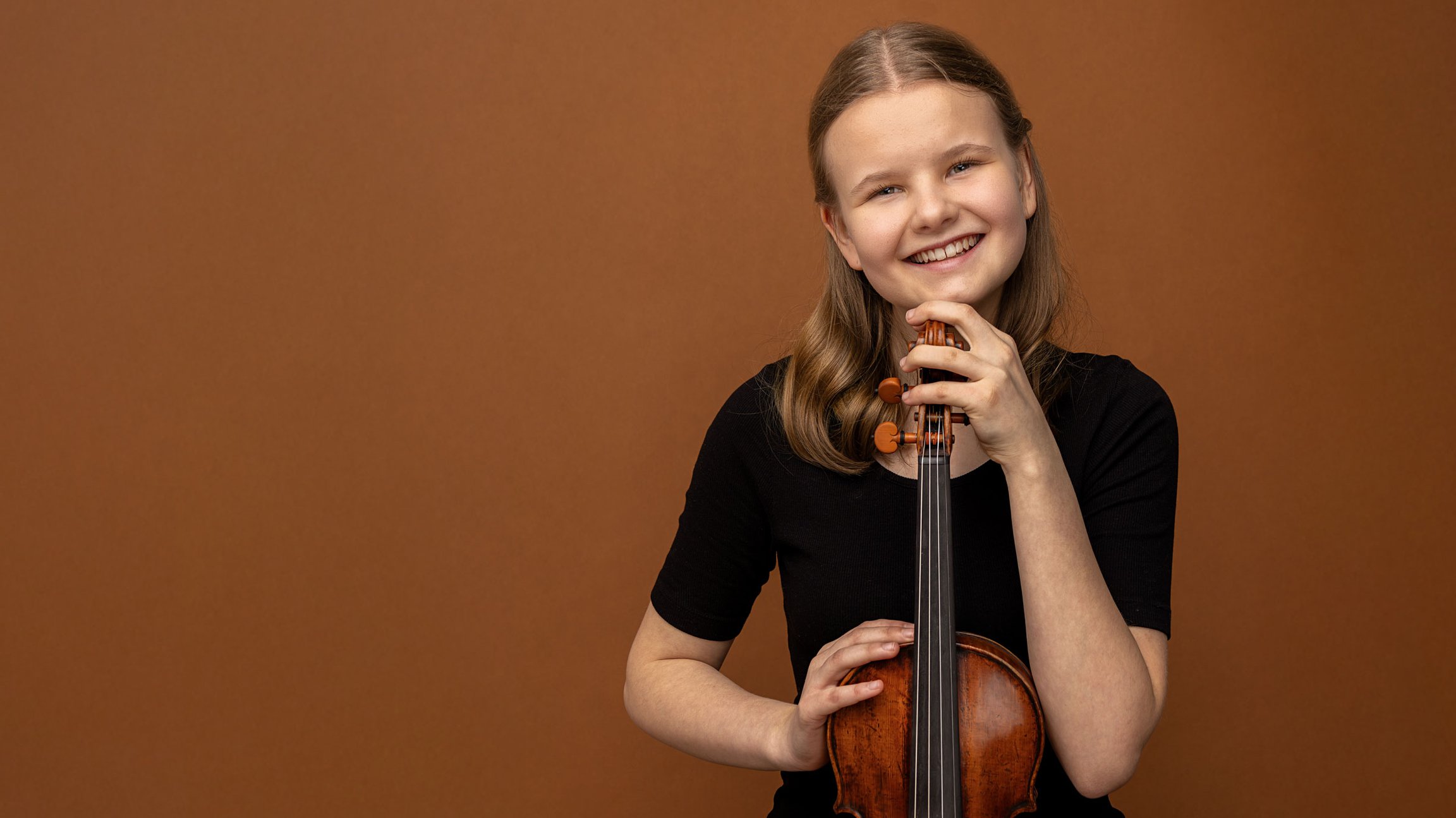 Eine jungen Person in schwarzem Oberteil vor warmbraunem Hintergrund, die eine Violine aufrecht hält und den Kopf auf den Wirbelkasten stützt, mit direktem Blick zur Kamera.