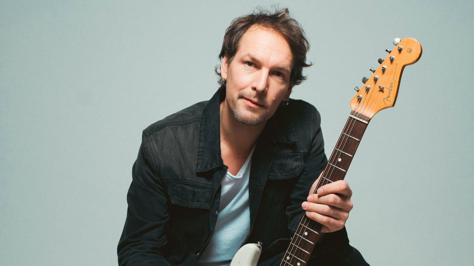 Person in dunkler Kleidung hält eine weiße E-Gitarre und kniet vor hellem Hintergrund. Studioaufnahme mit neutralem, grauem Hintergrund.