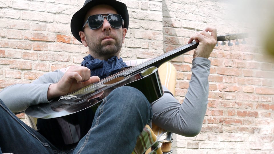 Foto für die Veranstaltung von Der Machatschek mit dem Künstler, der in einem Strandstuhl sitzt und eine Gitarre hält, und im Hintergrund eine Ziegelwand.