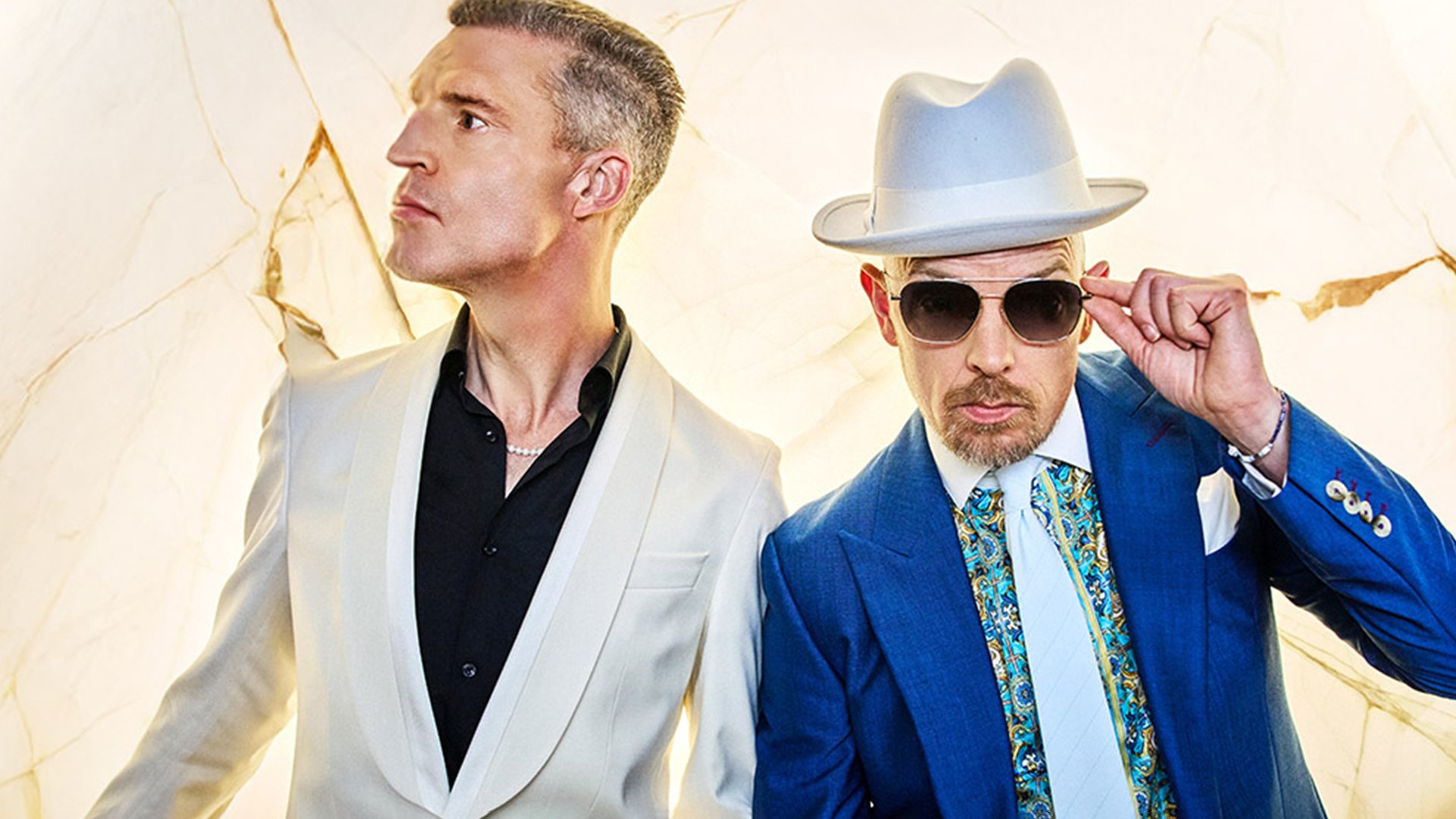 Foto für die Veranstaltung Benjamin von Stuckrad-Barre & Jan Delay – VORGLÜHEN 2026 mit den beiden Künstlern, von denen einer einen Hut und eine Sonnenbrille trägt, beide vor einem goldenen Hintergrund