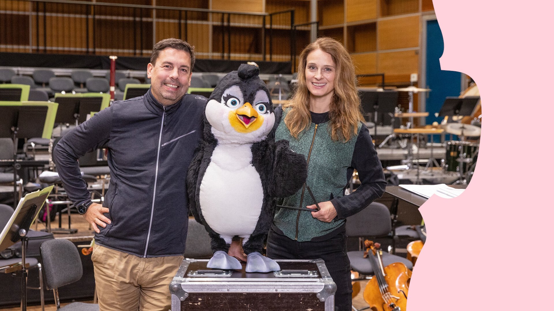 Zwei Personen und ein großes Stofftier, im Hintergrund ein Orchestersaal 