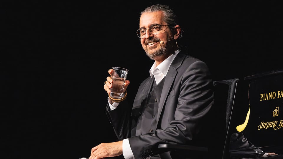 Bühnenfoto von Hagen Rether in dunklem Anzug mit Brille und Headsetmikrofon, sitzend neben einem Flügel und ein Wasserglas haltend, vor schwarzem Bühnenhintergrund.