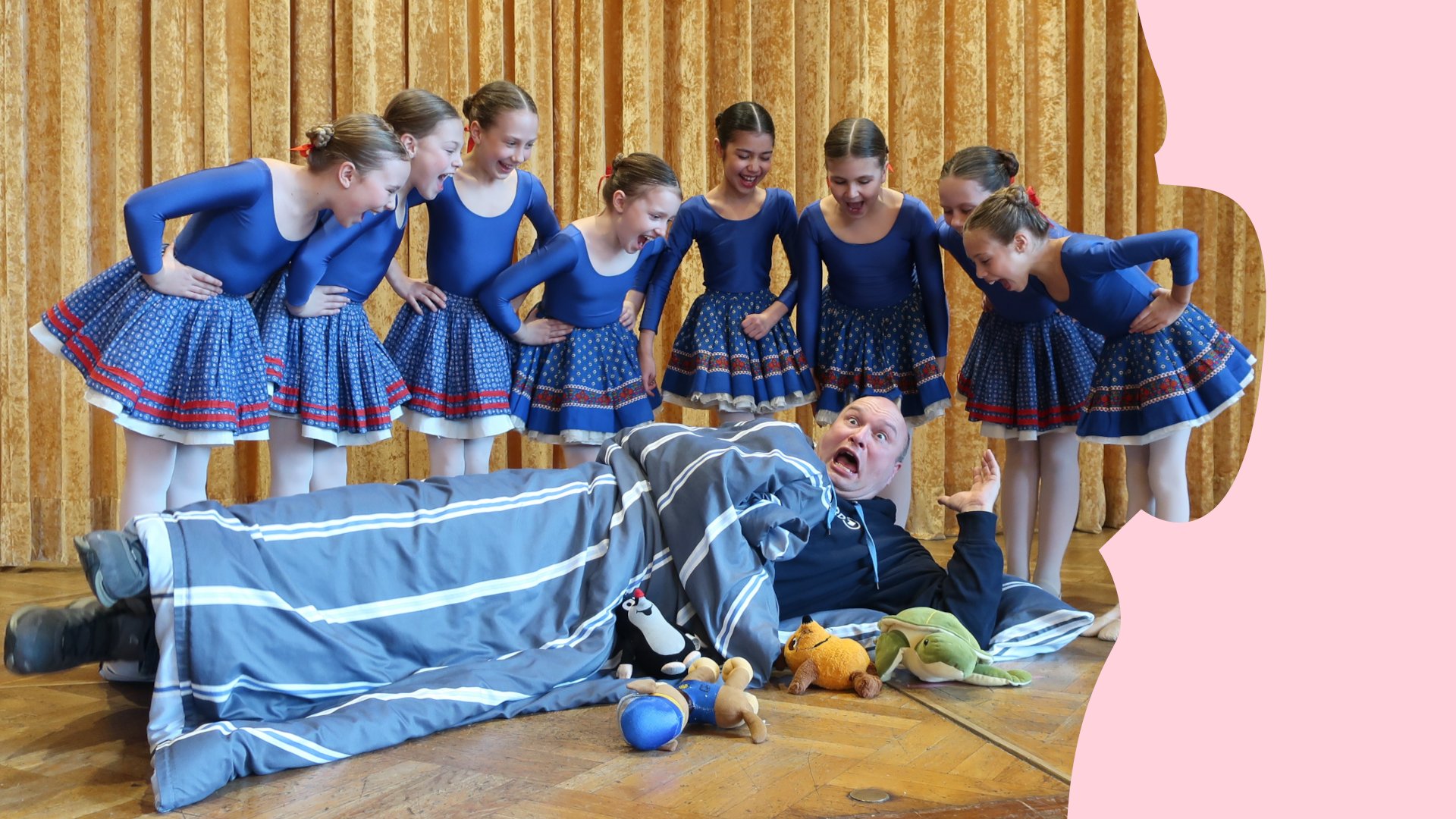 Gruppe von Kindern in blauen Tanzkostümen steht auf einer Bühne um eine Person, die zwischen Bettdecke und Stofftieren auf dem Boden liegt.