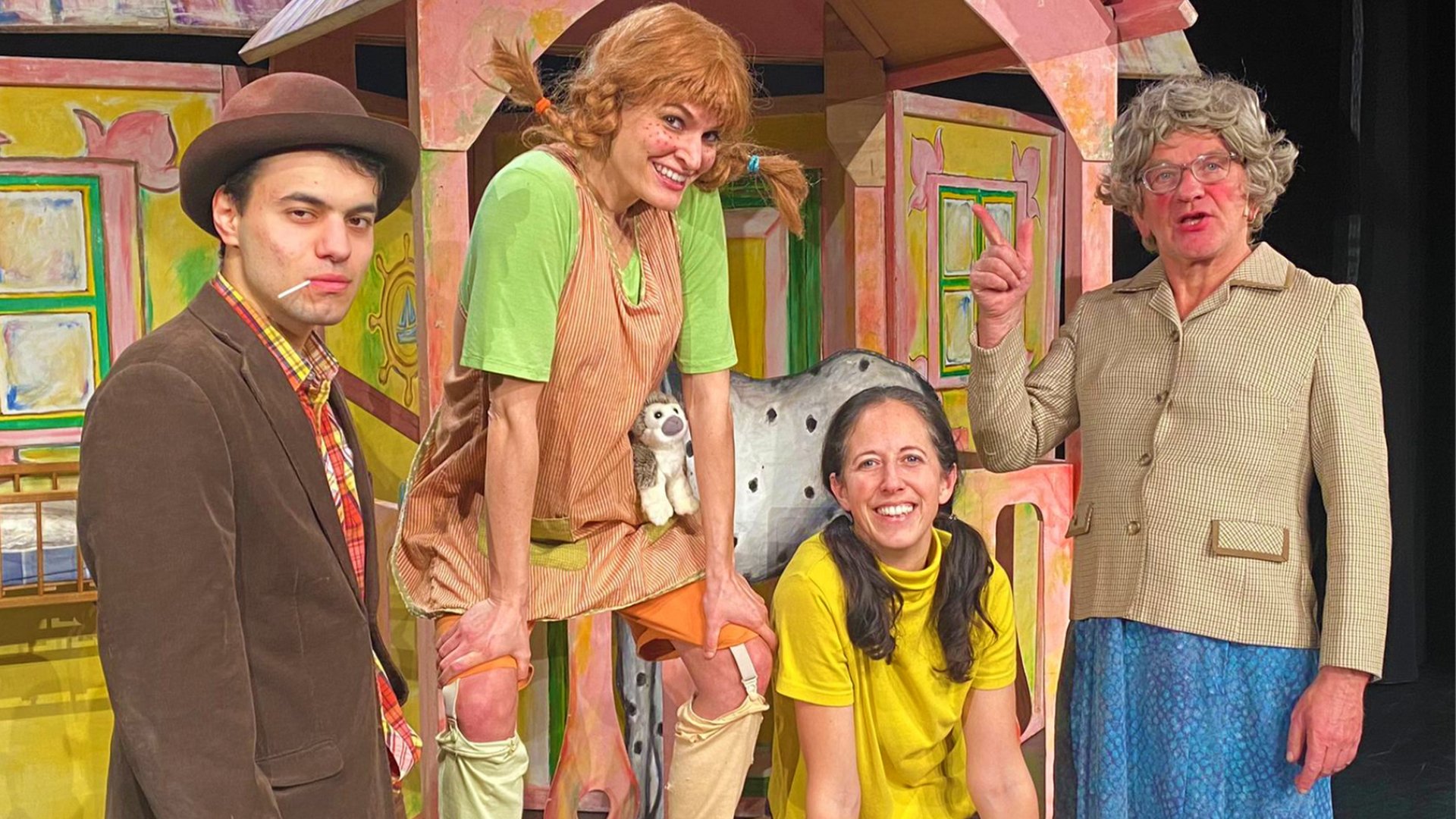 Vier Darsteller:innen in bunten Kostümen auf einer Theaterbühne vor einer farbig bemalten Holzkulisse mit Hausfassaden, Fenstern und schiefen Dächern; eine Figur ist als Pippi Langstrumpf verkleidet.