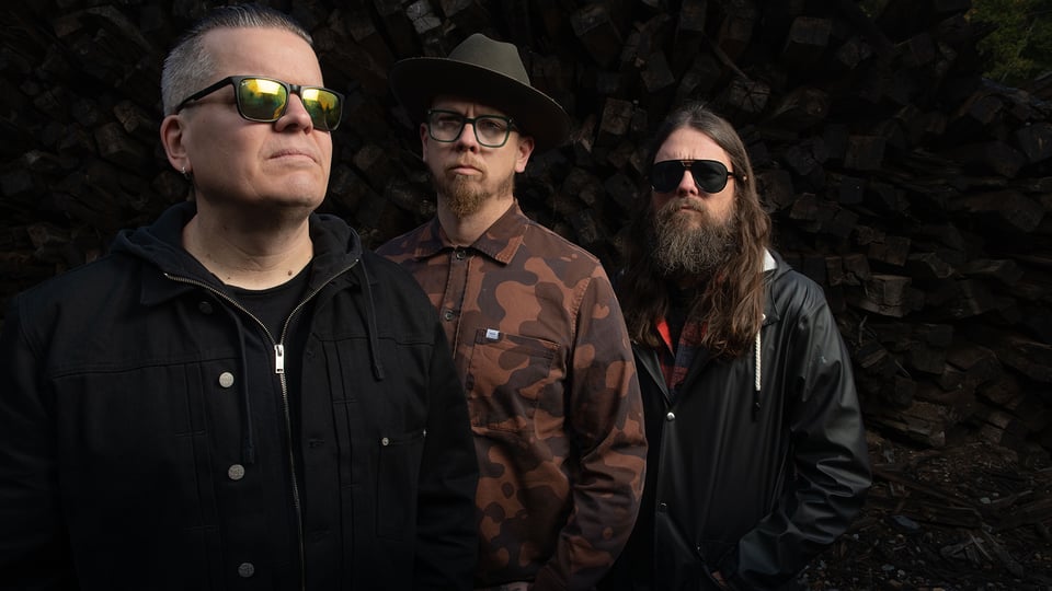 Monolord stehen nebeneinander vor einem dunklen Hintergrund aus gestapeltem Holz; sie tragen dunkle Kleidung, zwei mit Sonnenbrille, einer mit Hut, und blicken ernst in unterschiedliche Richtungen