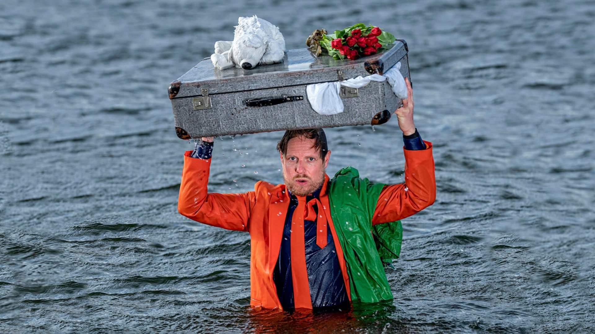 Pressefoto von Kabarettist Patrick Nederkoorn im Wasser stehend mit Koffer über dem Kopf.