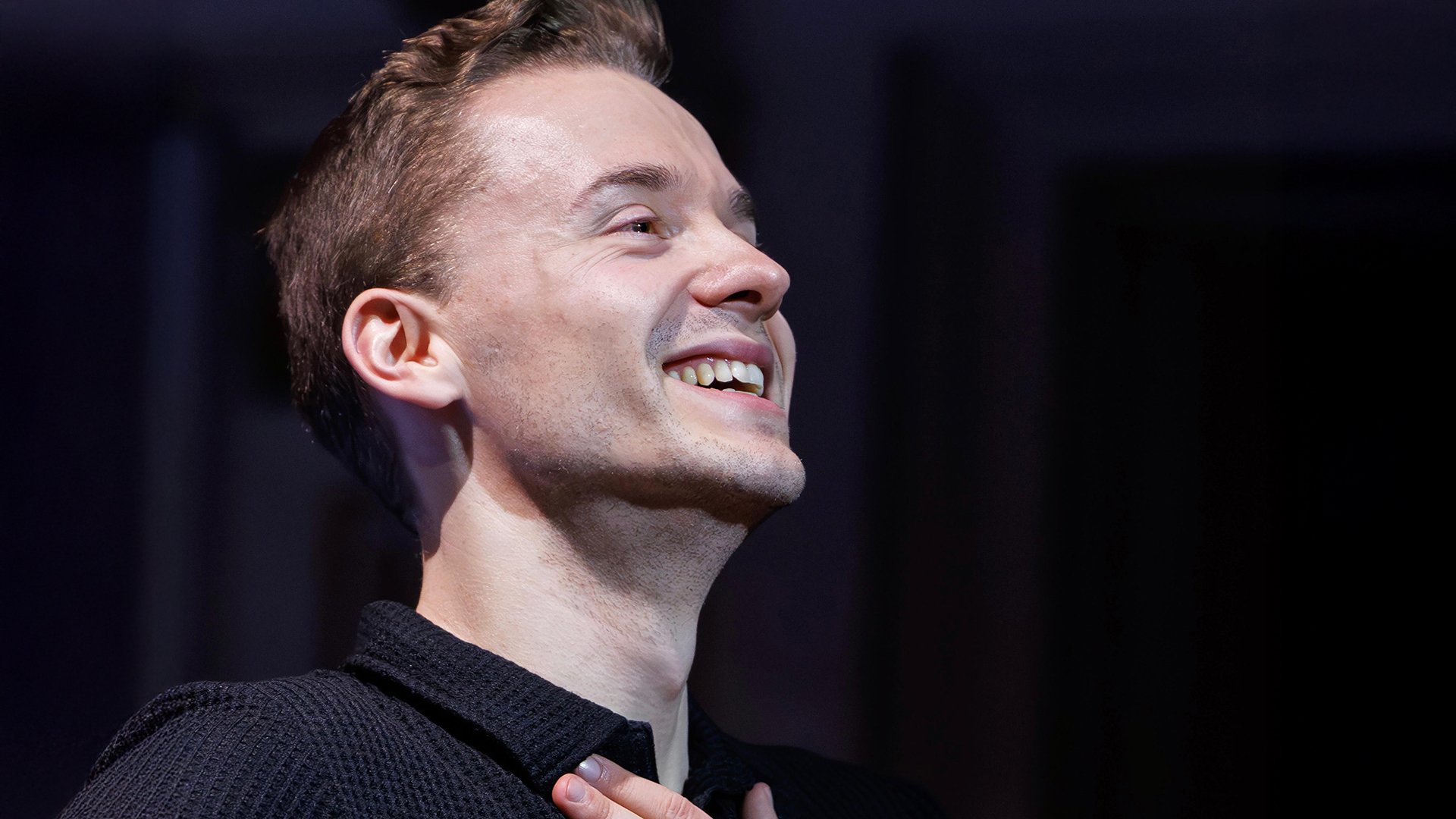 Porträt eines lächelnden Mannes auf einer Bühne, Blick nach oben, Hand auf der Brust, in stimmungsvoller Beleuchtung.